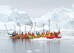 Kajakgruppe med pagajer i luften foran isbjerg. Photo by David D. Grant