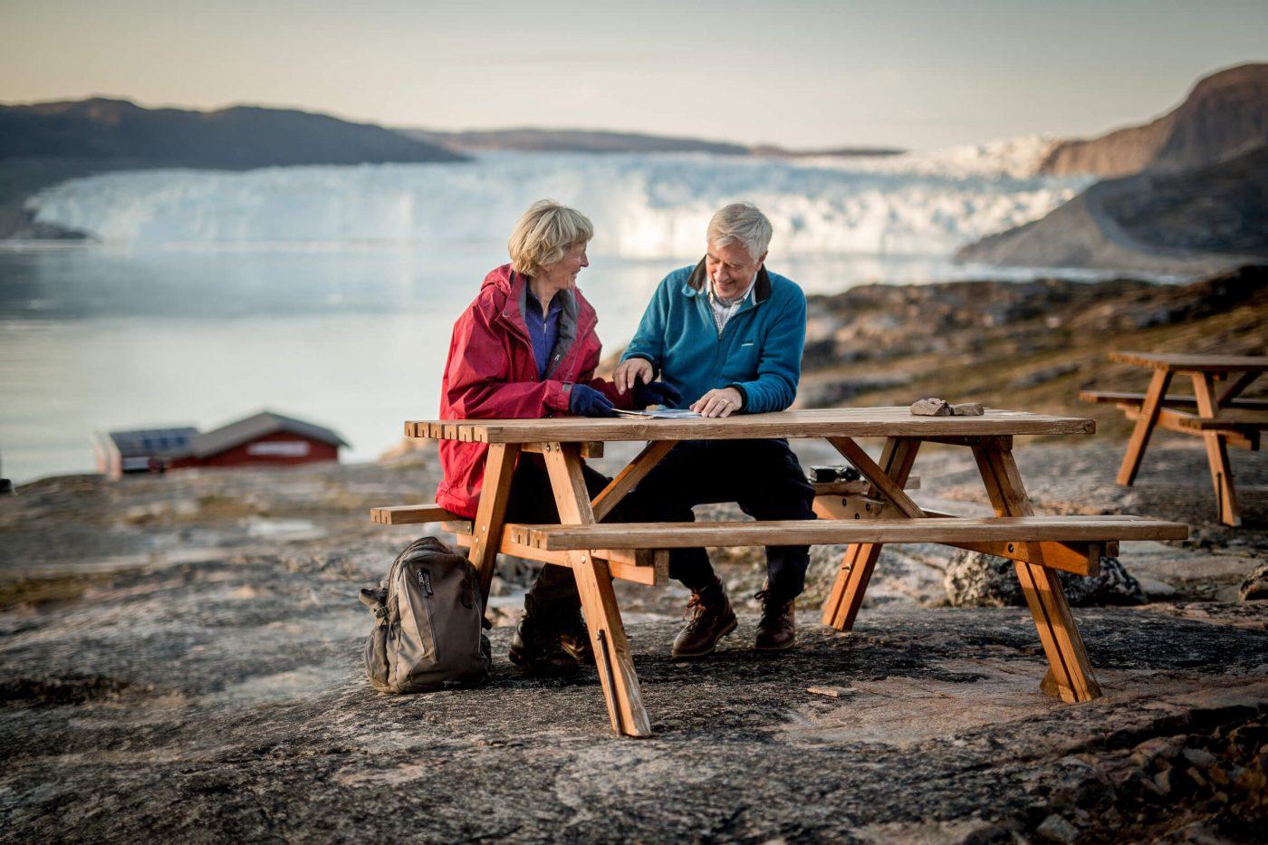 Two guests at Eqi Glacier Lodge in Greenland at a picnic table with the glacier in the background. Photo by Mads Pihl, Visit Greenland