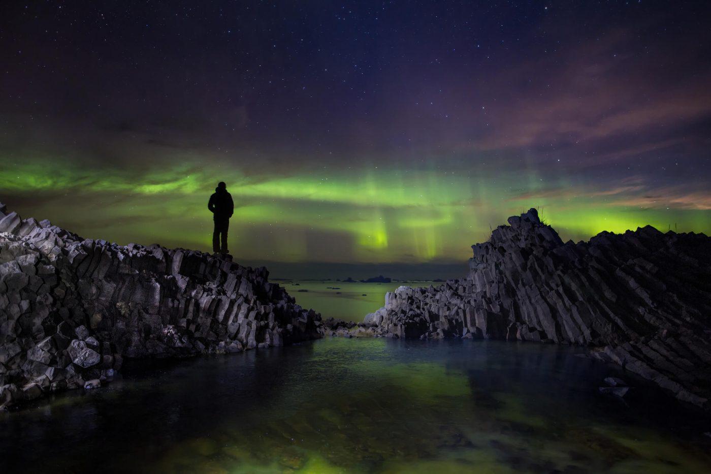 Hiker standing on volcanic rooks on Disko Island in North Greenland looking at northern lights dancing over the ocean. Photo by Paul Zizka