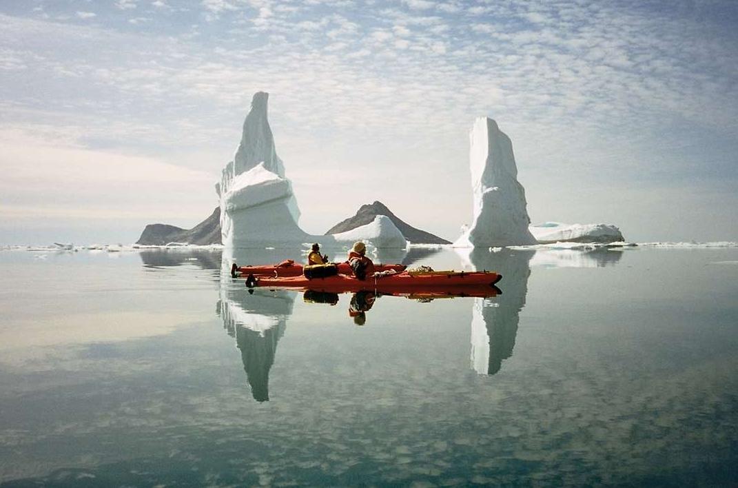 Two people kayaking next to large icebergs in crystal clear water. Photo by Arctic Dream, Visit Greenland