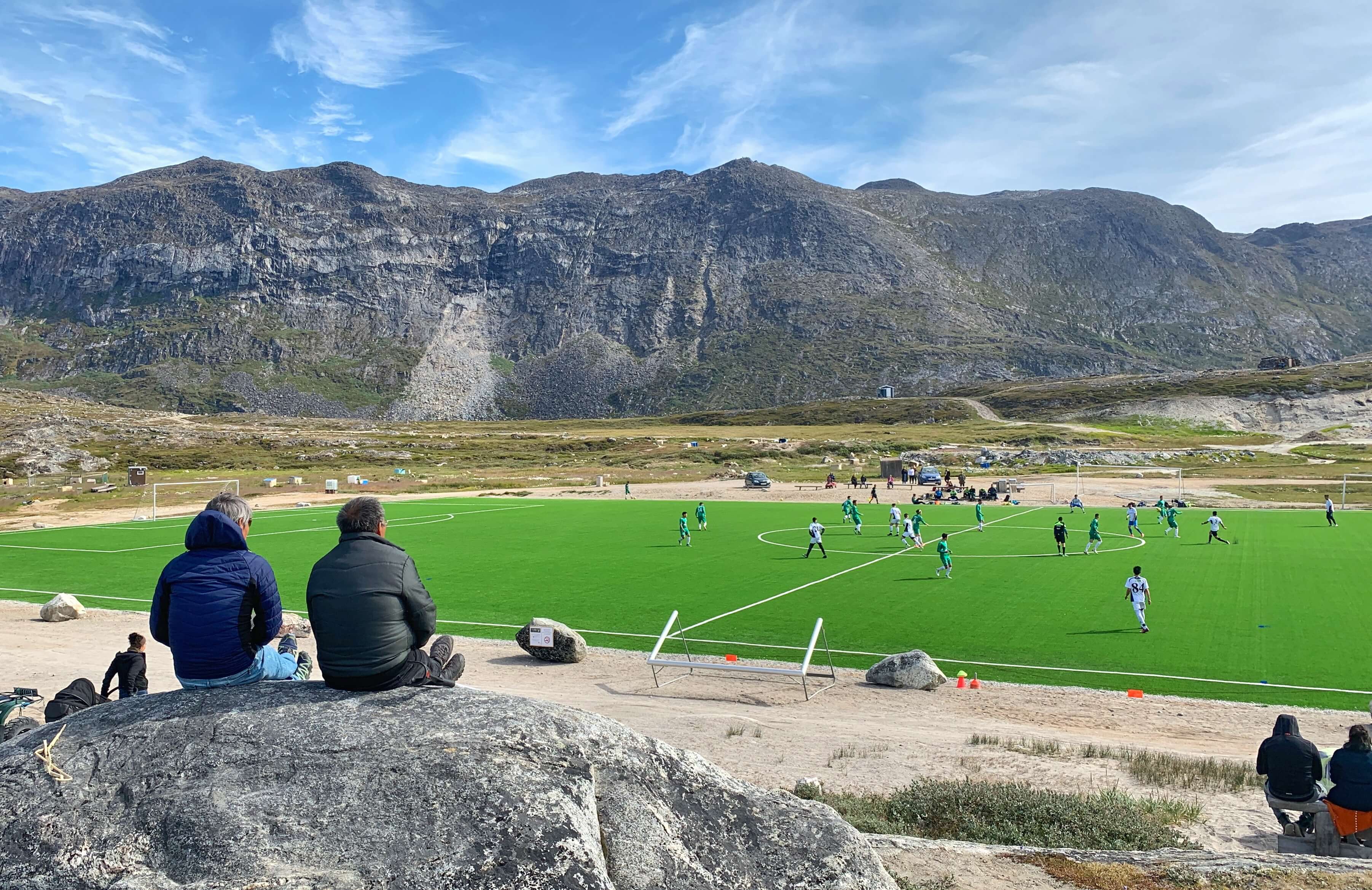 Two people sitting on a rock watching a soccer game. Photo by Espen Andersen, Visit Greenland