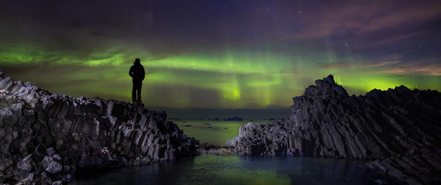Hiker standing on volcanic rooks on Disko Island in North Greenland looking at northern lights dancing over the ocean. Photo by Paul Zizka