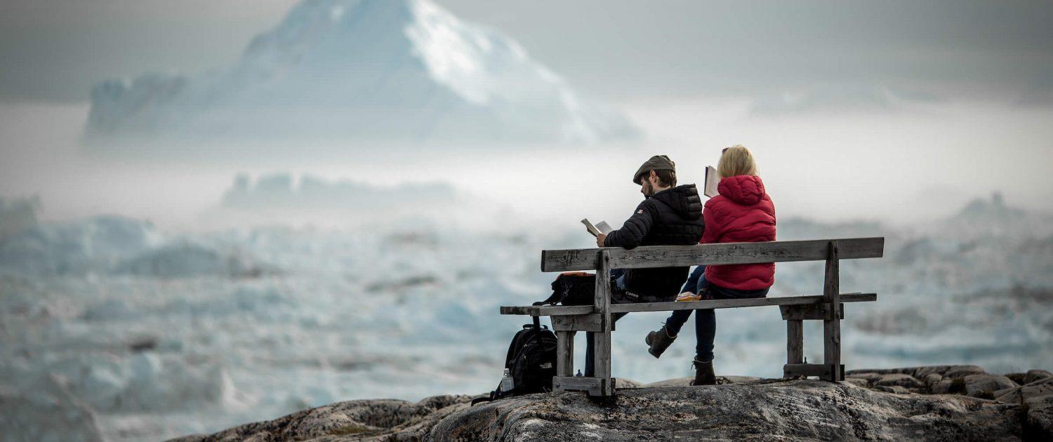 Two travelers reading on a bench overlooking icebergs in the Ilulissat ice fjord in Greenland. Photo by Mads Pihl