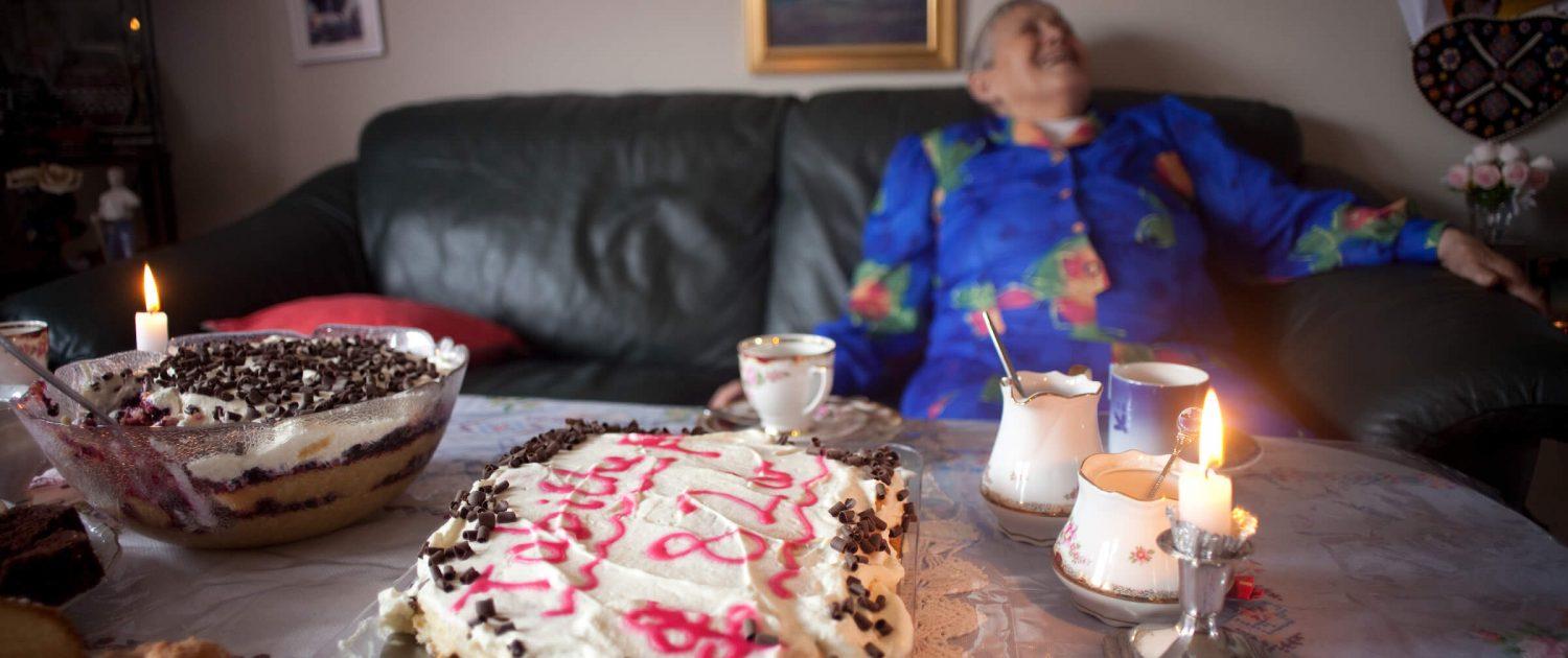 An elderly woman at a kaffemik laughing and enjoying herself. By Angu Motzfeldt