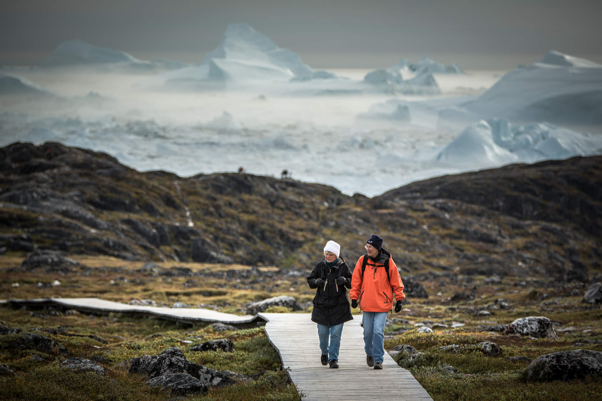Two women on the boardwalk near Sermermiut with icebergs in the background in Ilulissat in Greenland. Photo by Mads Pihl