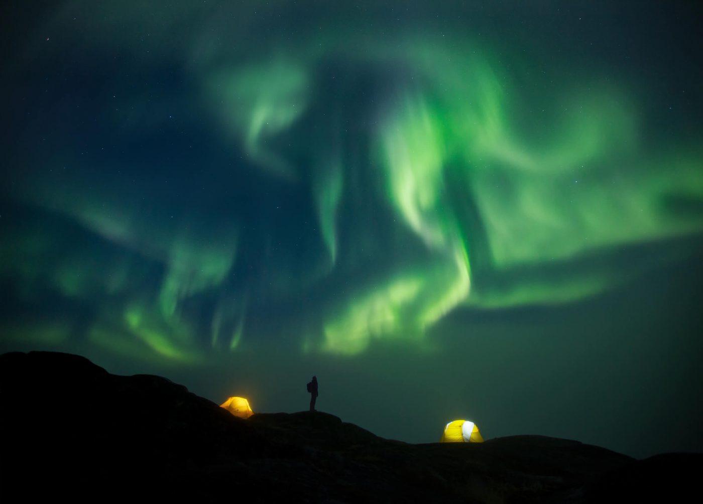 A happy backpacker stands outside her tent in her backcountry camp to watch the northern lights swirl over the Ilulissat Icefjord just beyond the hill. By Paul Zizka
