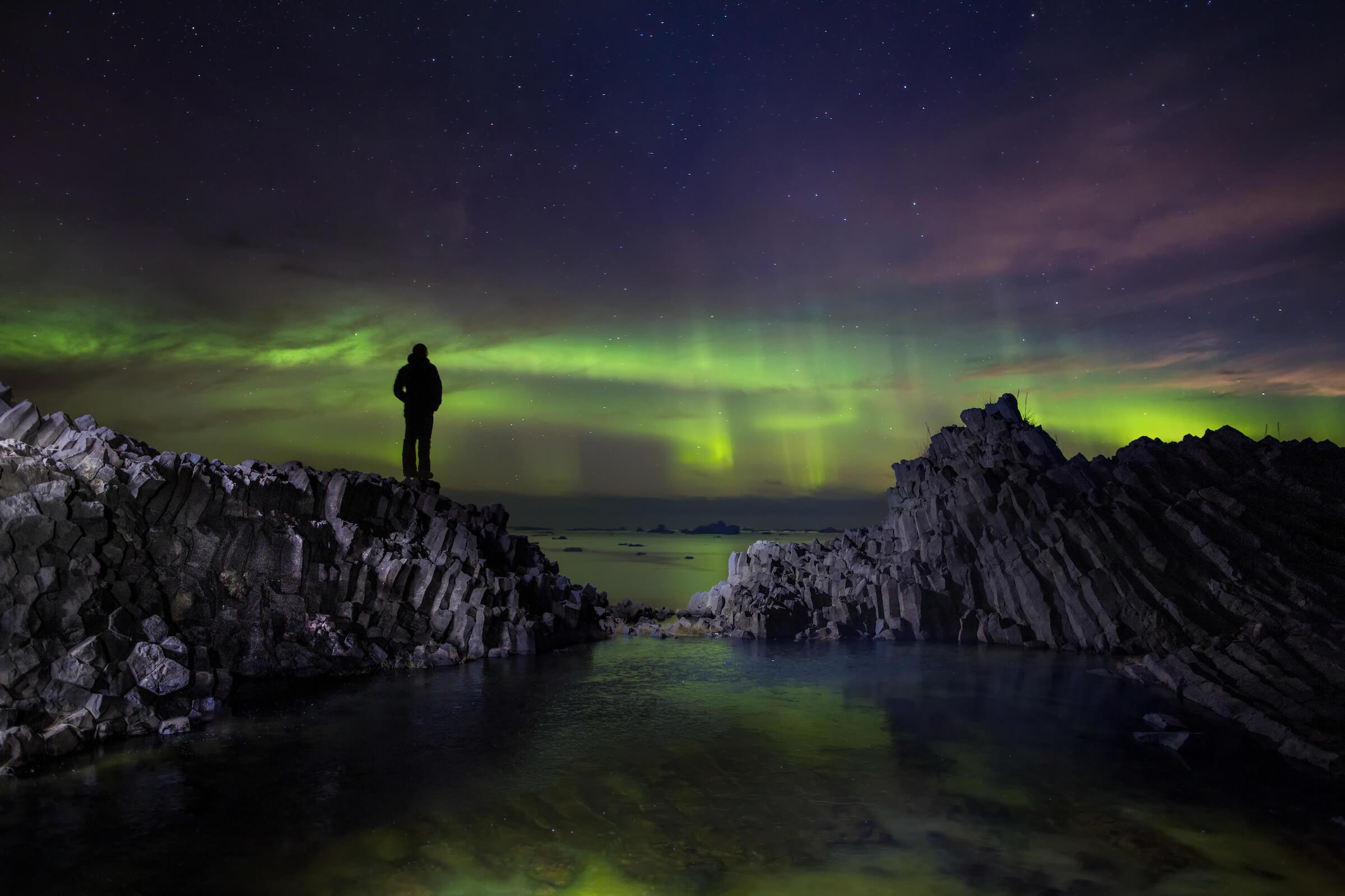 Hiker standing on volcanic rocks on Disko Island in North Greenland looking at northern lights dancing over the ocean. Photo by Paul Zizka - Visit Greenland