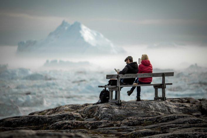 Two travelers reading on a bench overlooking icebergs in the Ilulissat ice fjord in Greenland. Photo by Mads Pihl